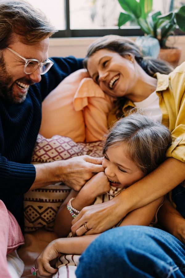 A happy family of three laughs and cuddles together on a couch surrounded by pillows and plants.