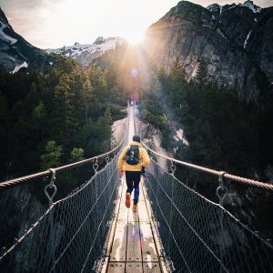 A person in a yellow jacket crosses a suspension bridge over a gorge with mountains in the background at sunrise.